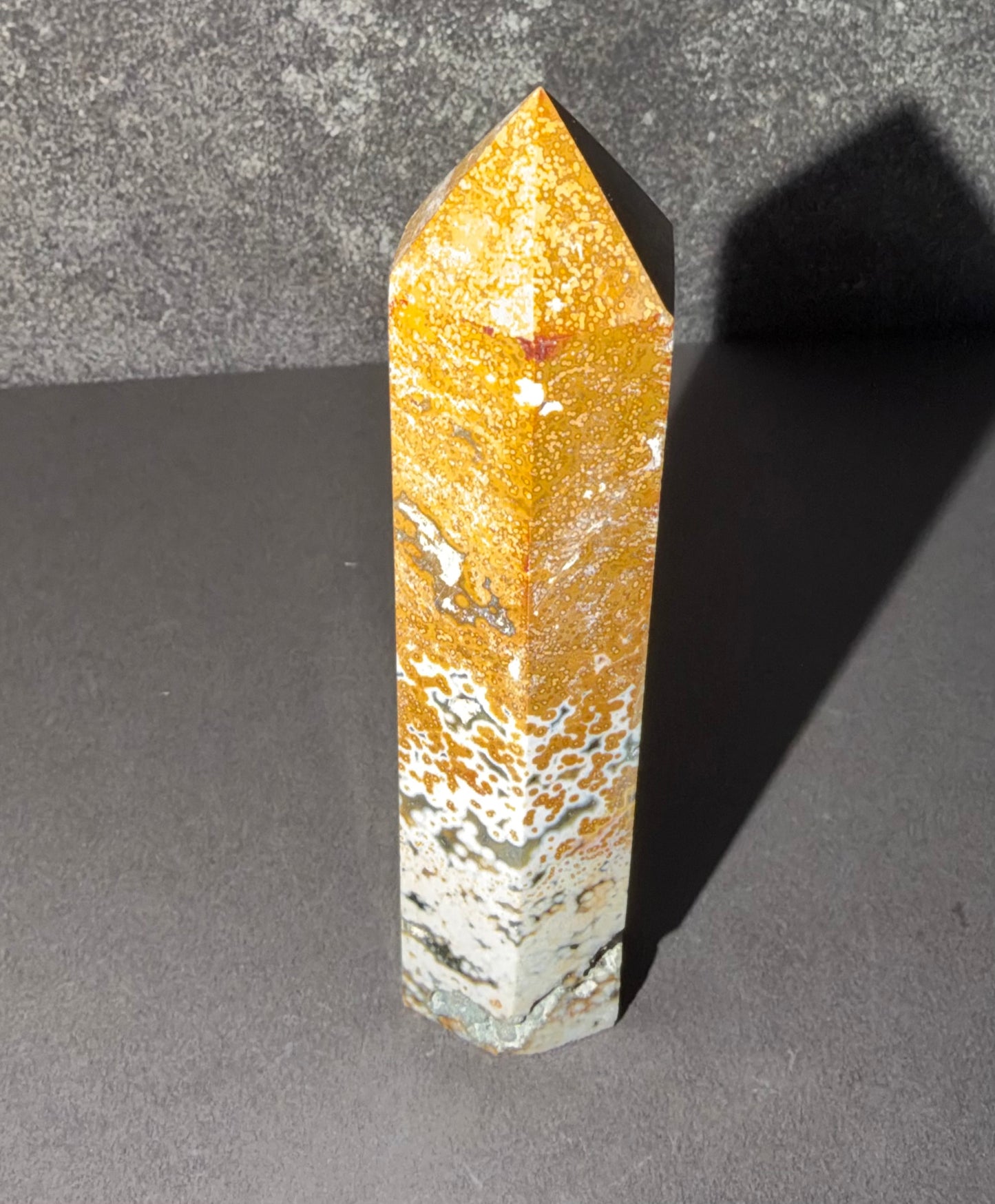 A polished crystal tower with a pointed top, featuring a marbled pattern of golden orange, white, and gray, standing on a dark gray surface with a gray textured background.