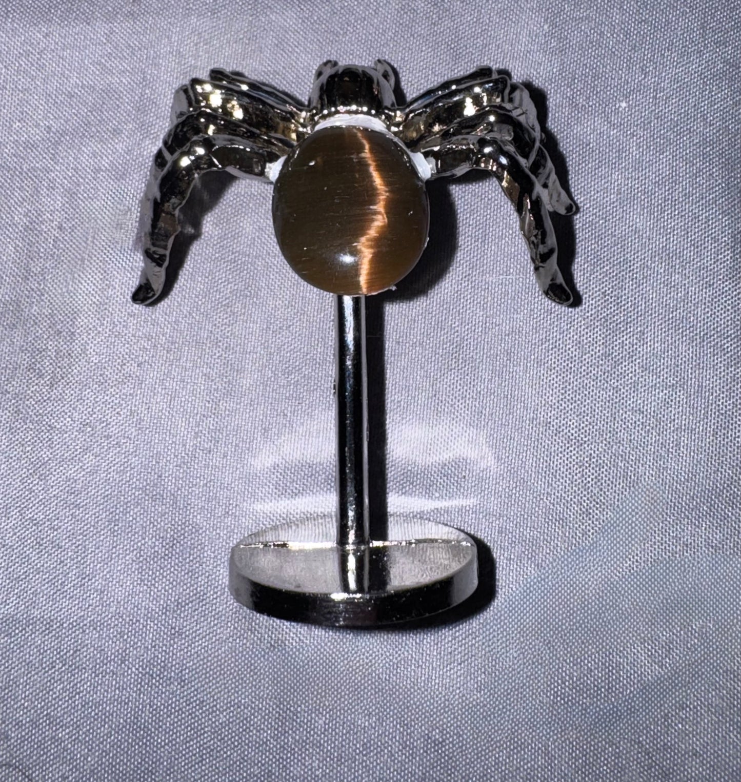 A spider-shaped stud earring with a polished silver-tone metal body and a tiger's eye stone in the center, displayed on a stand against a textured gray fabric background.