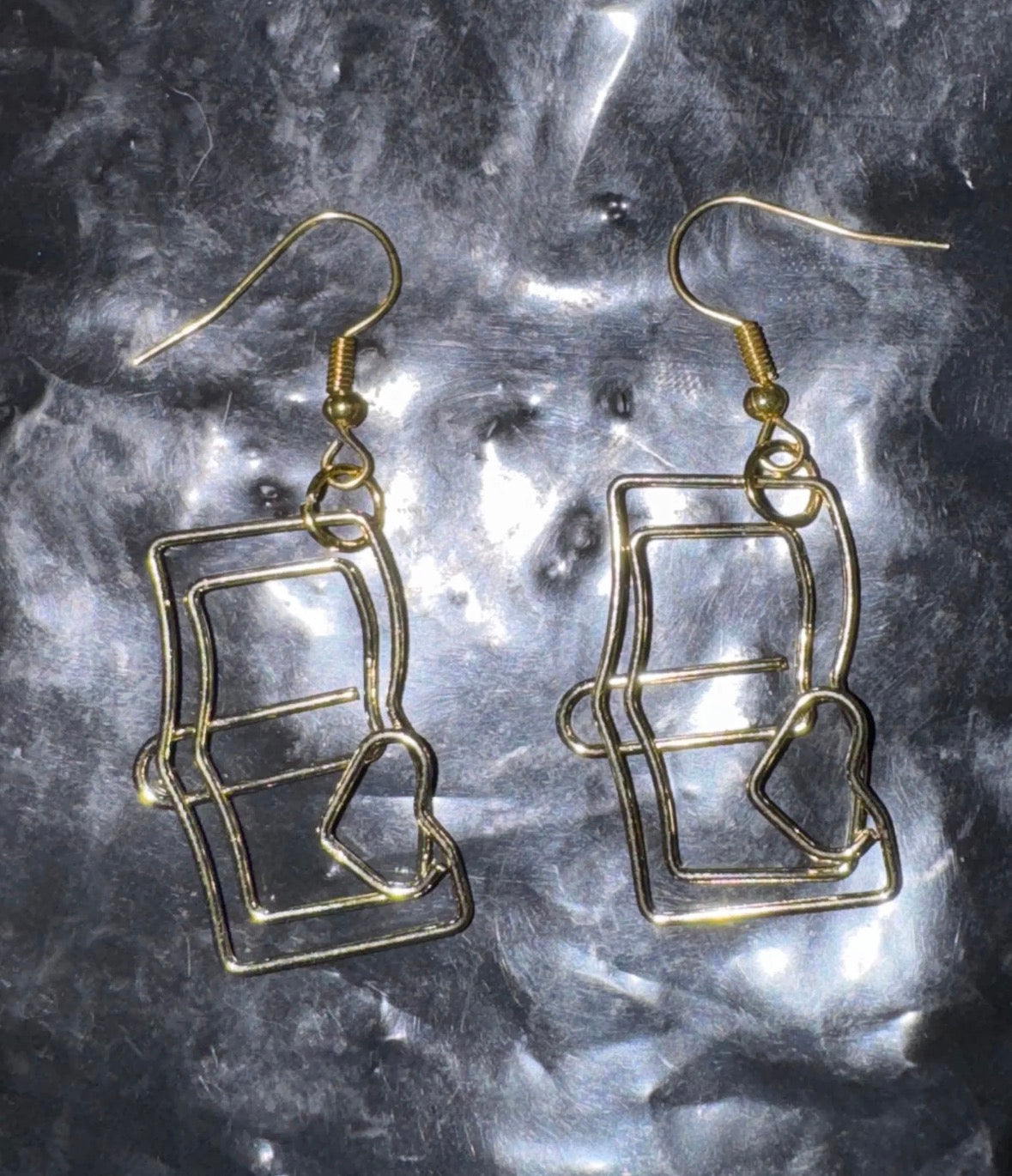 Gold wireframe book-shaped earrings on a reflective gray surface