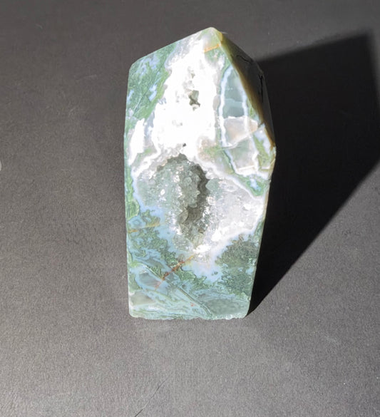 A polished natural crystal point featuring a green and white marbled pattern with a central white quartz cavity, photographed on a dark gray surface with a hard shadow.