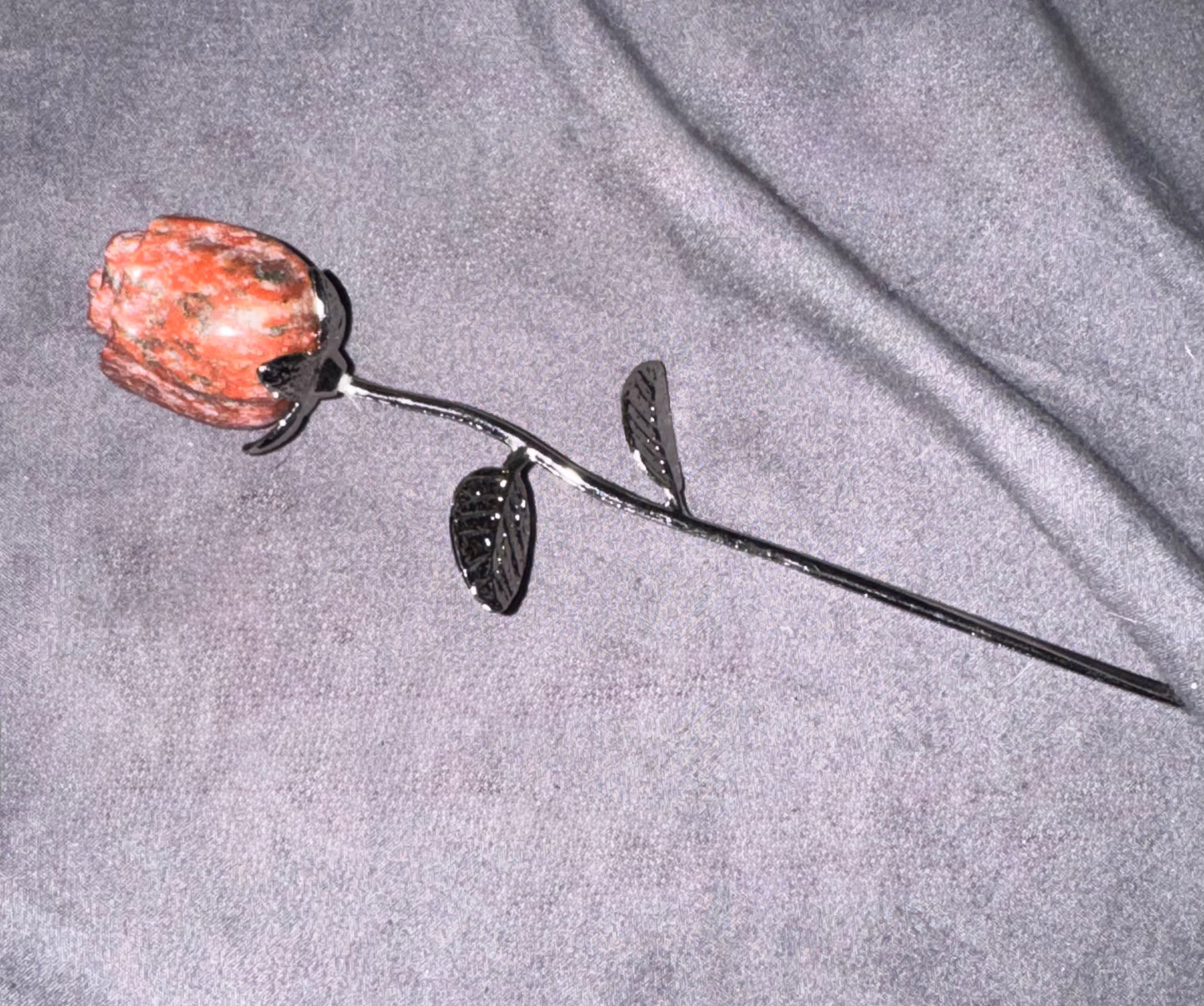 A handmade metal rose with a textured orange-red stone flower head and dark metal stem, leaves, and thorns, displayed on a gray fabric background.
