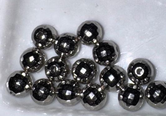 A group of faceted metallic silver beads with a dark center, arranged in a pile on a white textured background.