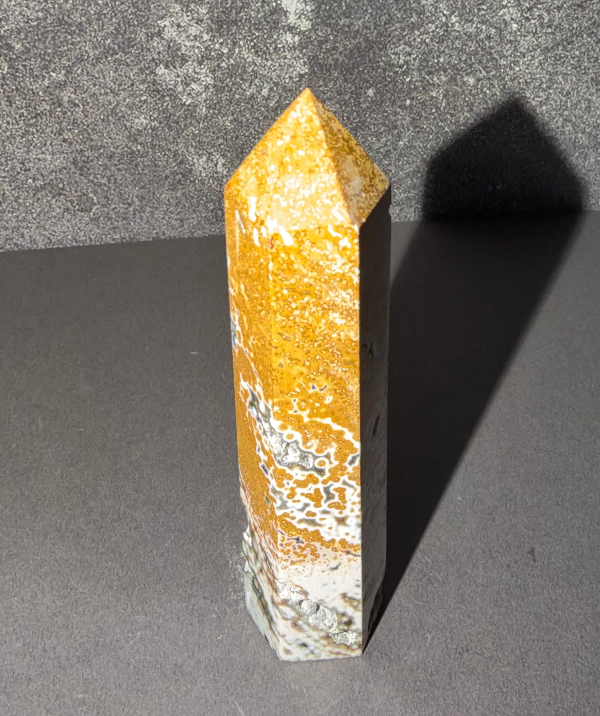 A polished golden-yellow and white agate crystal obelisk standing upright on a dark gray surface, casting a sharp shadow against a textured gray background.