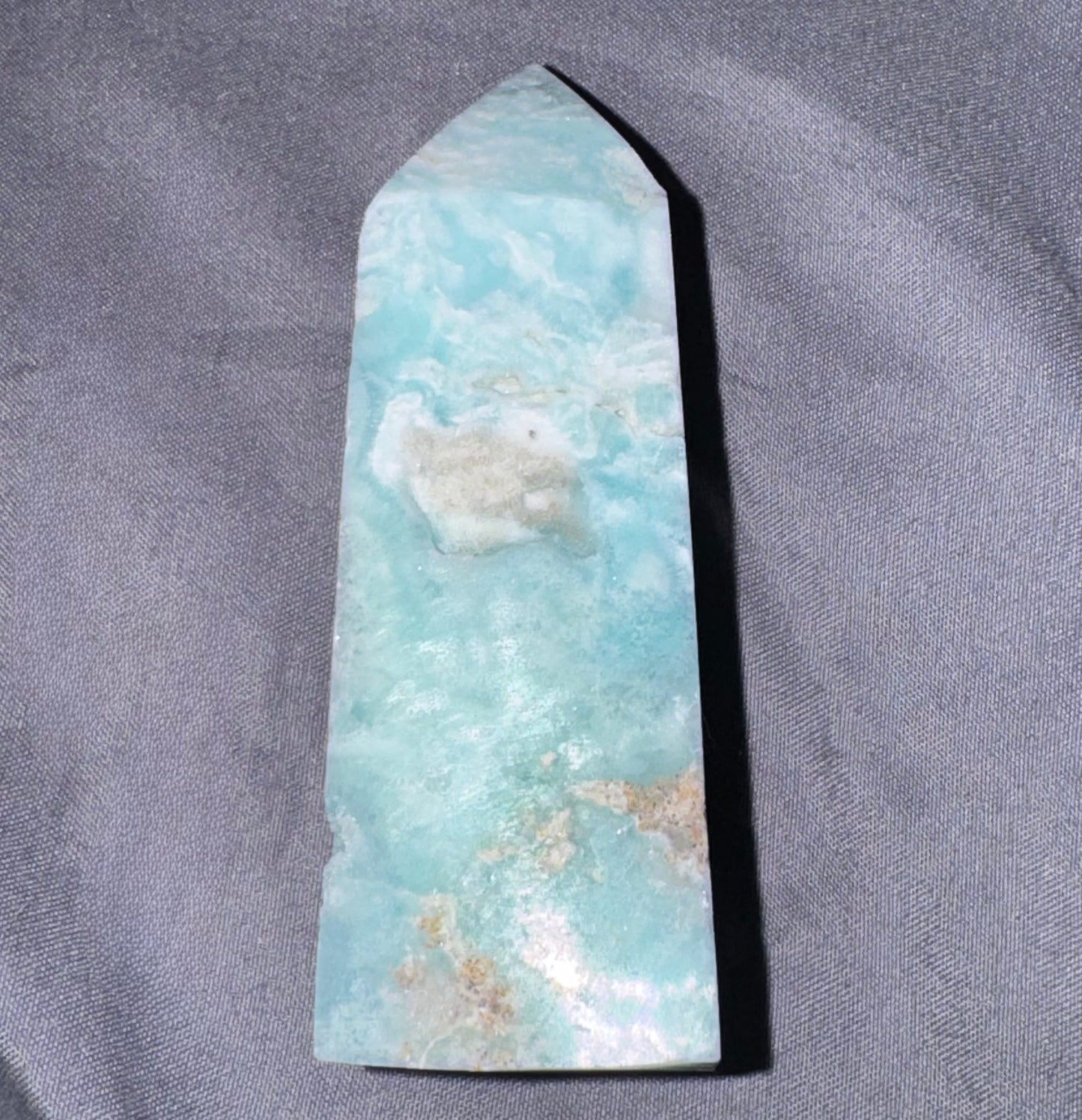 A polished  crystal point, featuring natural light blue and white marbled patterns with small tan inclusions, displayed on a textured gray fabric background.