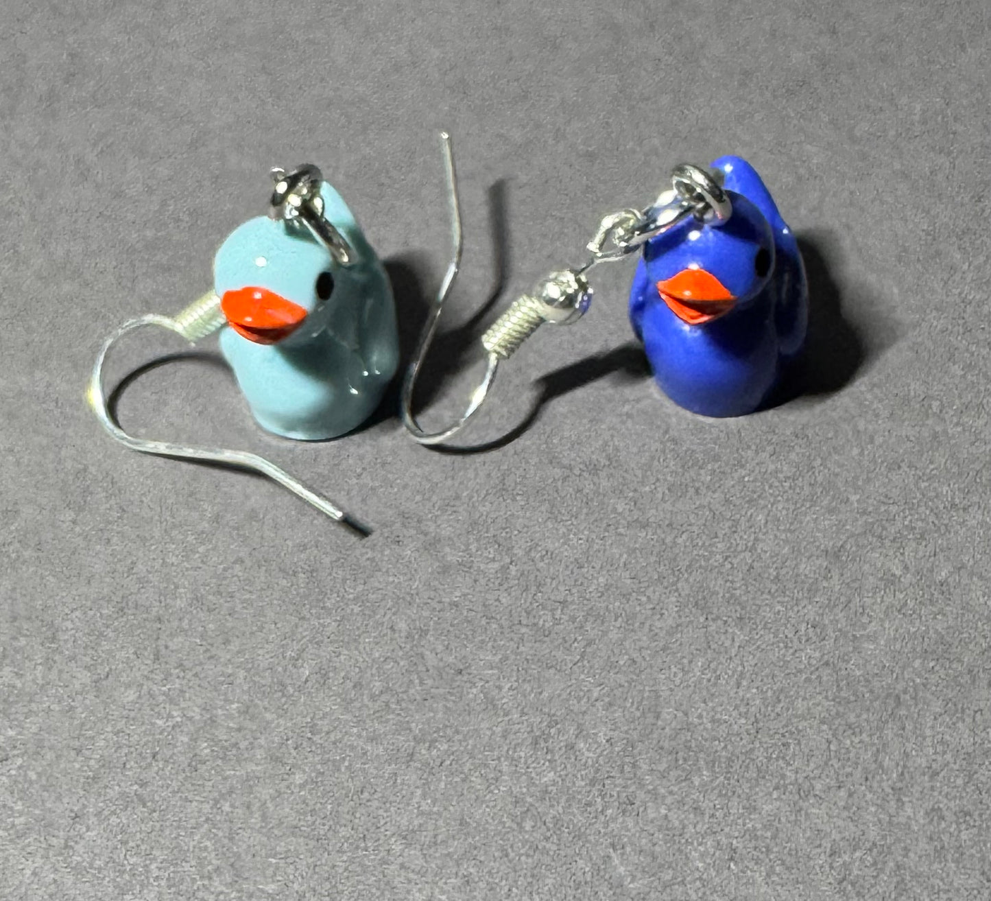 A pair of small duck-shaped dangle earrings on a gray textured background. One earring features a light blue duck with an orange beak and silver fishhook ear wires, and the other features a dark blue duck with an orange beak and silver fishhook ear wires.
