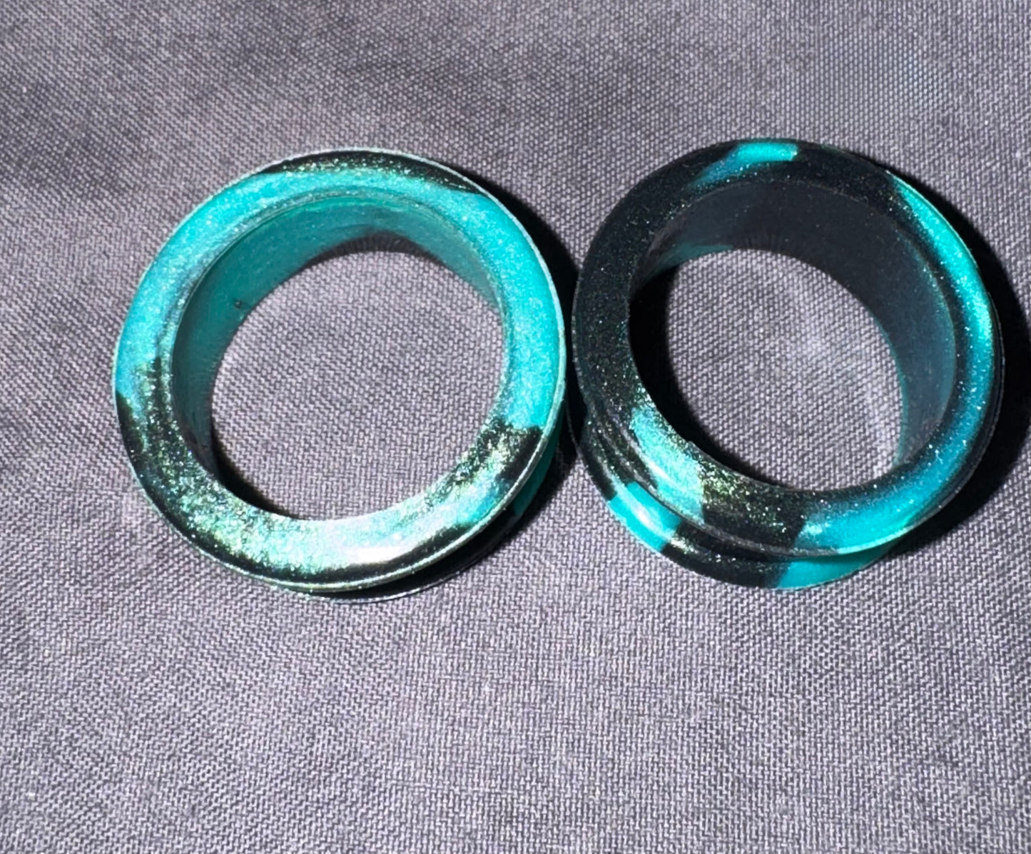 Two circular turquoise and black rings with a glittery finish, displayed on a gray textured fabric background.