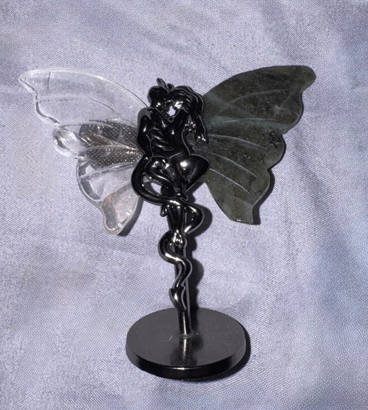 A black  figurine with one clear transparent wing and one dark gray wing, mounted on a round black base against a light gray fabric background.