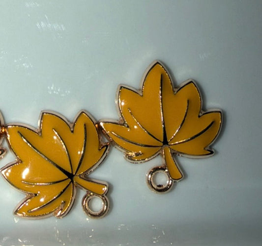 A pair of yellow maple leaf charms with gold-tone metal borders and hanging loops, displayed on a light blue surface.