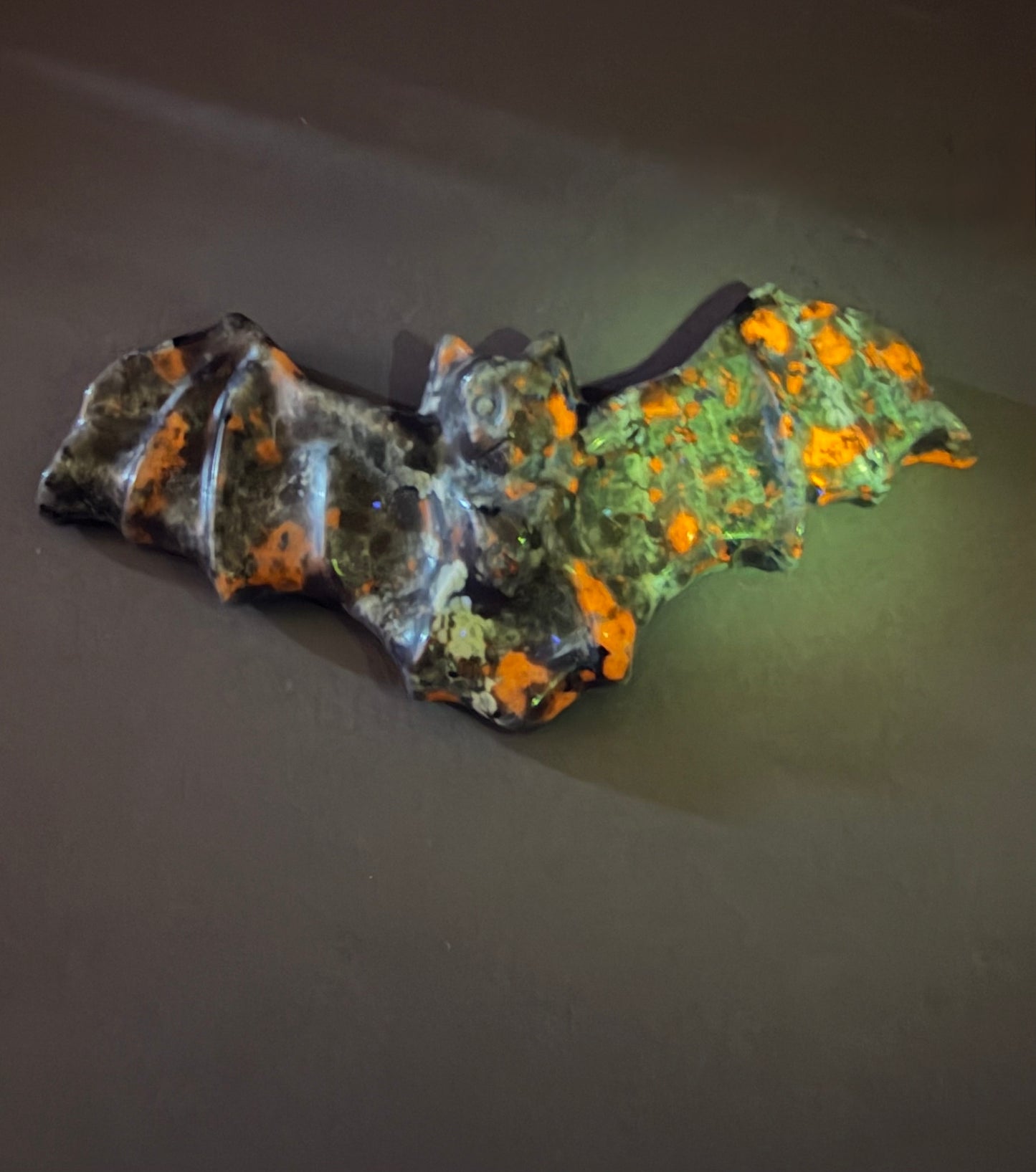 A carved bat figurine made from dark stone with bright orange and green mineral inclusions, photographed on a dark gray surface.