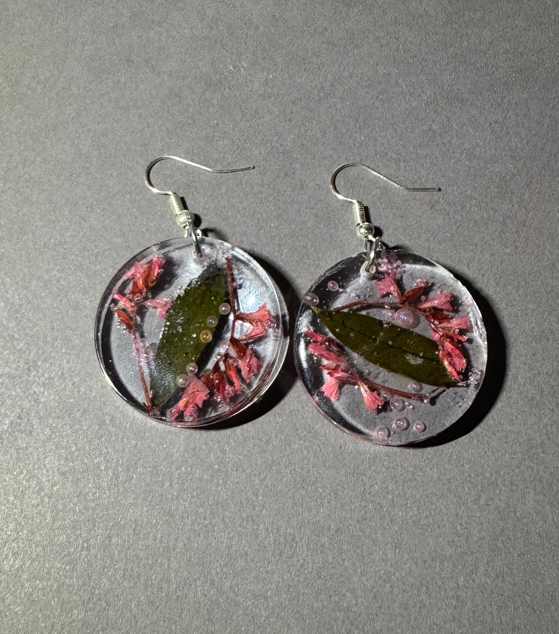 A pair of round clear resin drop earrings with silver fishhook wires, containing dried green leaves and pink sea plants, displayed on a textured gray background.
