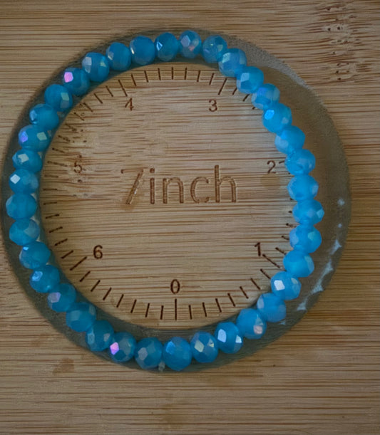 A beaded bracelet made of faceted light blue beads, shown in a circular arrangement on a wooden measuring board marked with inch measurements and the word 'Zinch'.