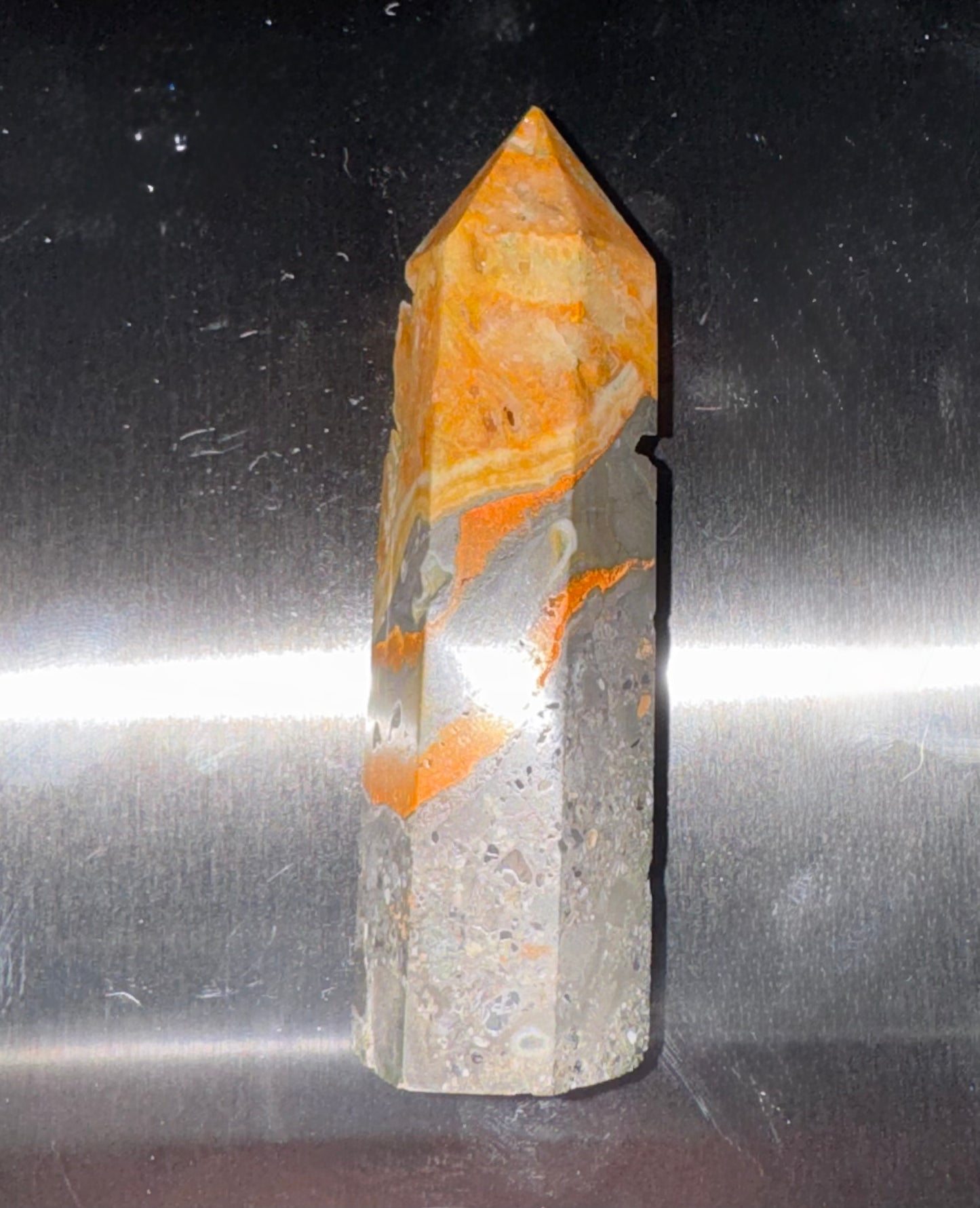 A natural quartz crystal point featuring vibrant orange and yellow banded patterns over a translucent gray body, standing upright on a reflective metallic surface.