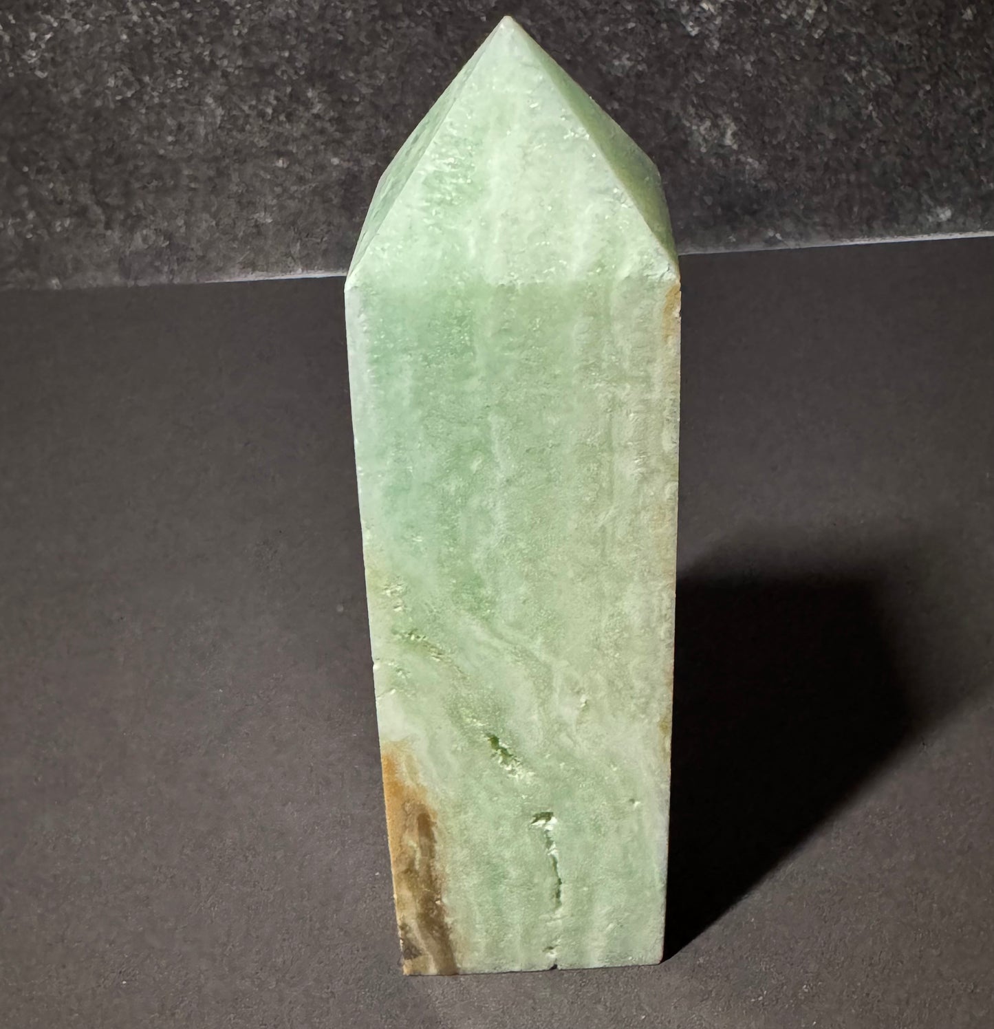 A polished light green crystal tower or obelisk with natural marbling and brownish inclusions near the base, standing upright on a dark surface against a dark gray background.