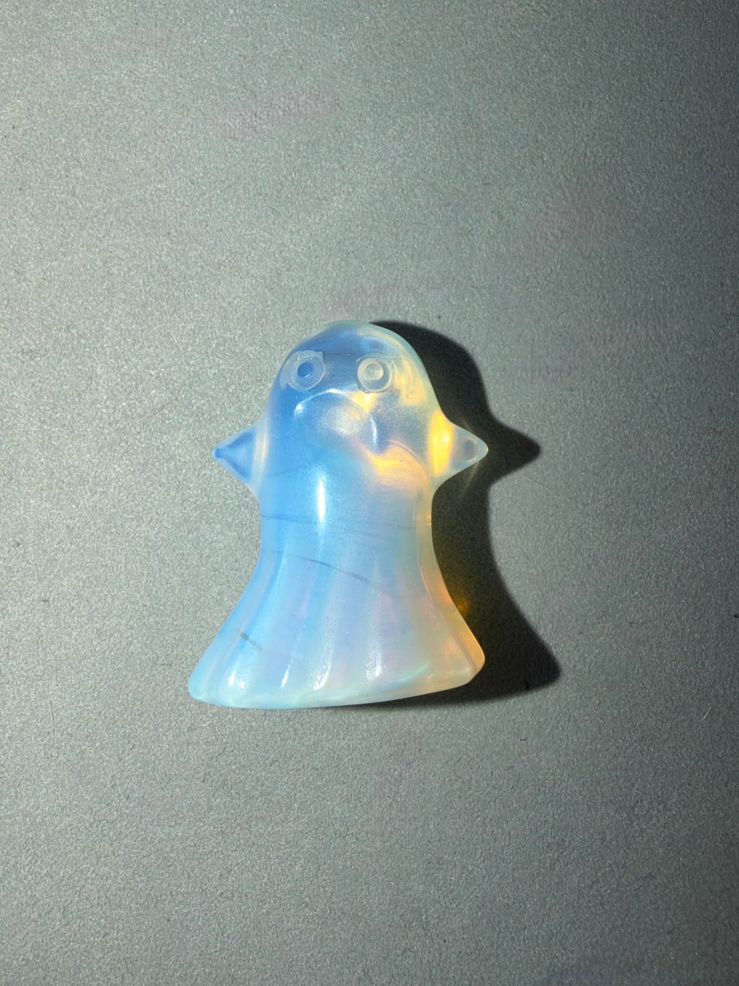 A small ghost-shaped pendant made of iridescent opal, showing shifting pastel blue, yellow, and white colors, photographed on a plain gray surface with a hard shadow.
