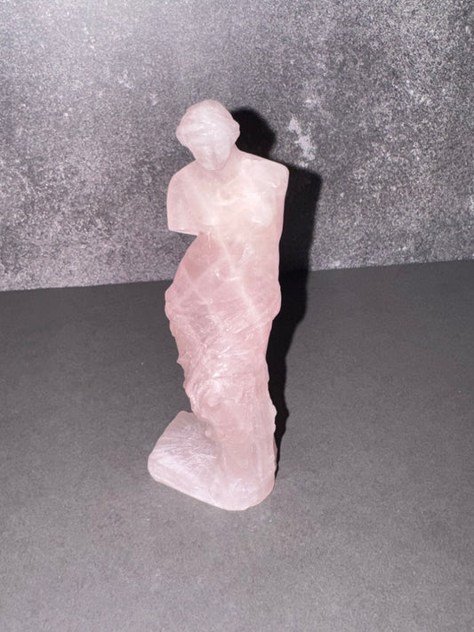 A small pink stone figurine of a draped female statue, photographed against a dark gray textured backdrop on a gray surface.