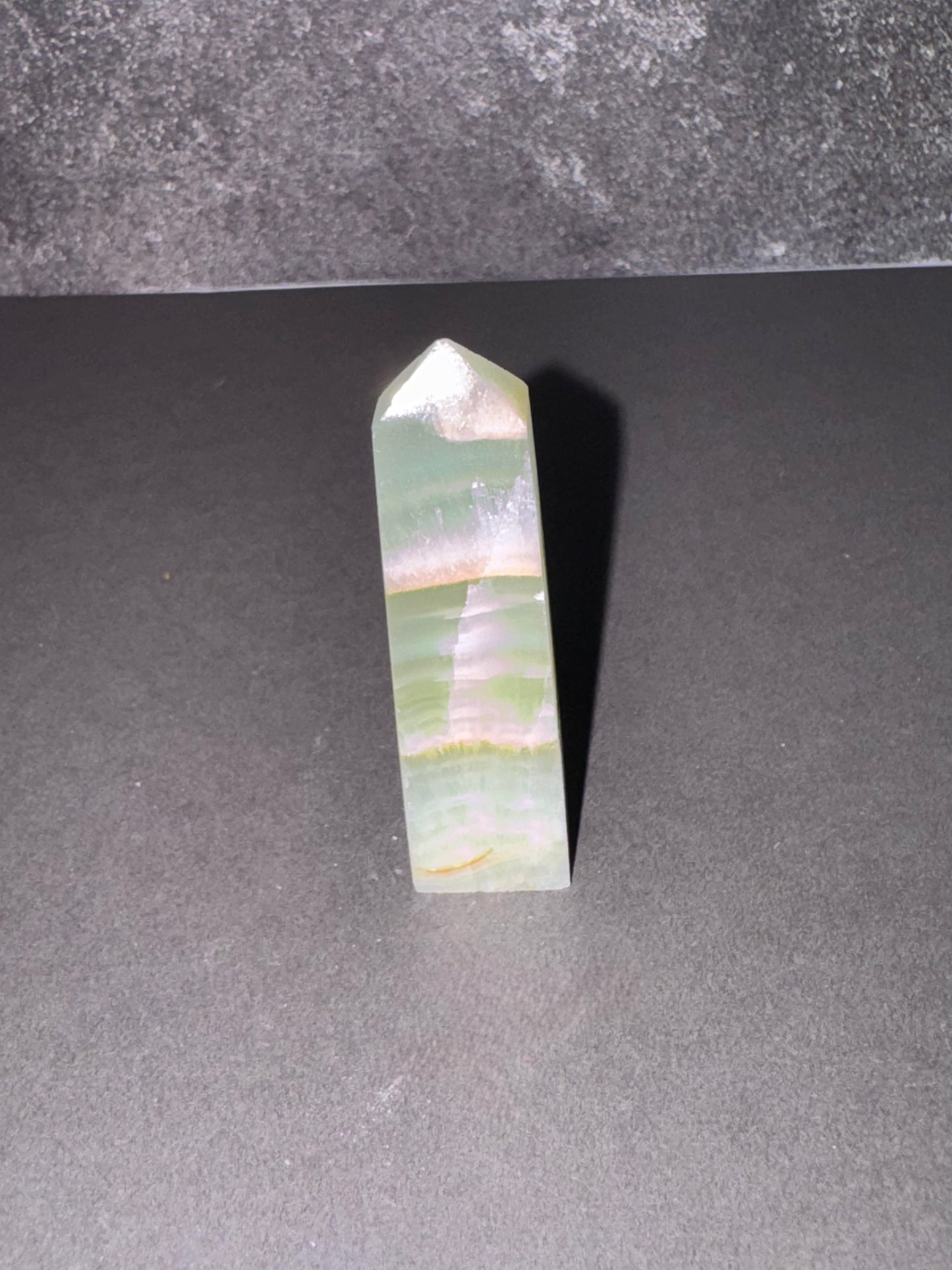 A natural green aventurine crystal point with horizontal banding, standing upright on a dark gray surface against a textured gray background.
