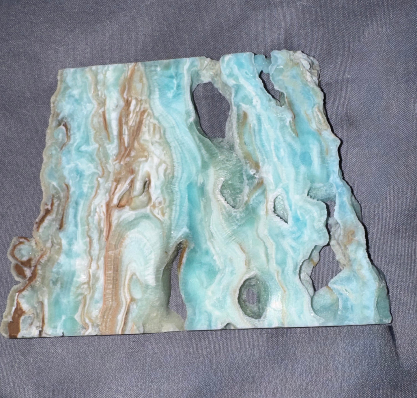 A polished stone slab with organic, irregular edges, featuring swirling patterns of light blue, turquoise, cream, and tan, displayed on a textured gray fabric background.