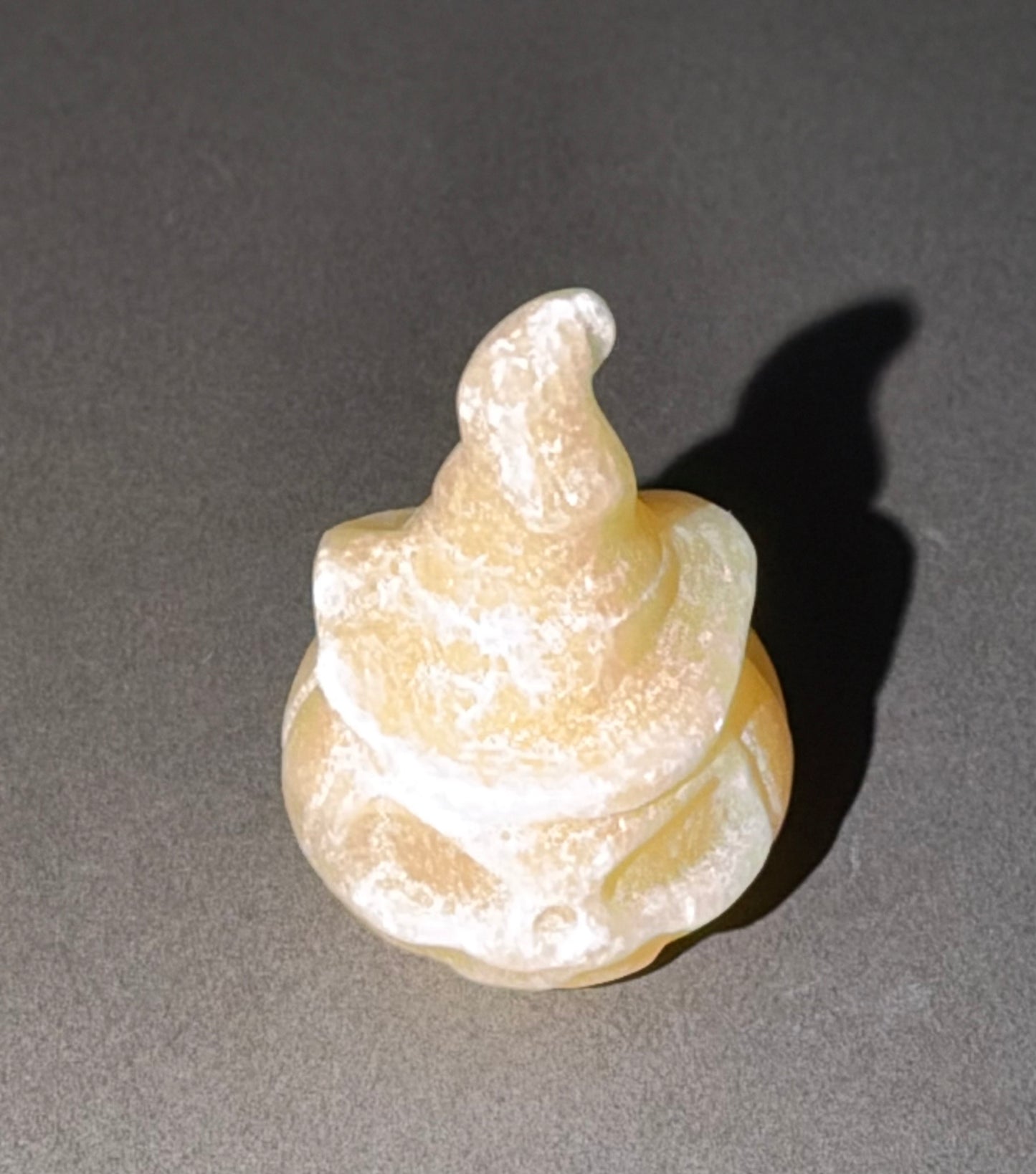 A small sculpted figurine resembling a coiled, textured spiral shape in a pale yellowish-beige translucent material, photographed on a dark gray surface with a sharp shadow.