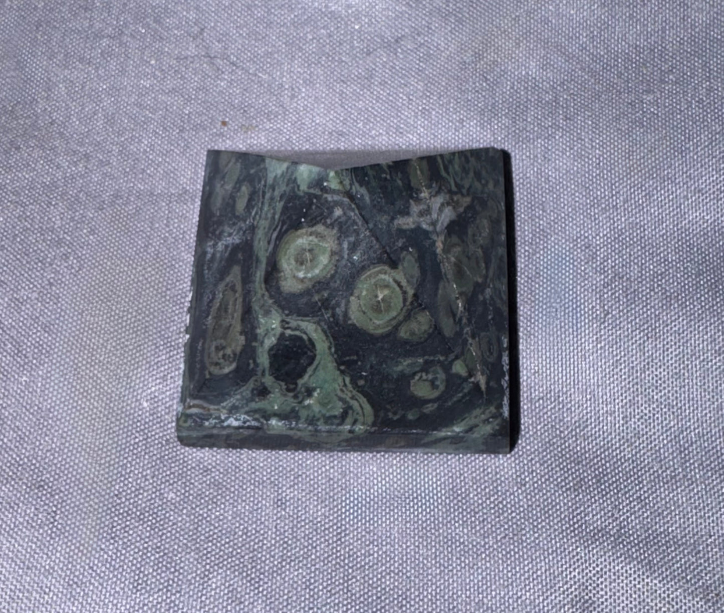 A rectangular polished stone slab with a marbled pattern of dark green, black, and light olive green, resting on a textured gray fabric background.