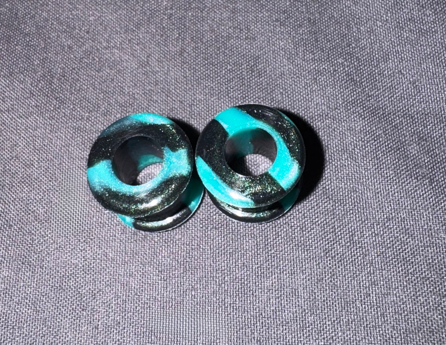 Two round glass plug earrings with a marbled pattern of turquoise, black, and fine gold glitter, displayed on a textured gray fabric background.