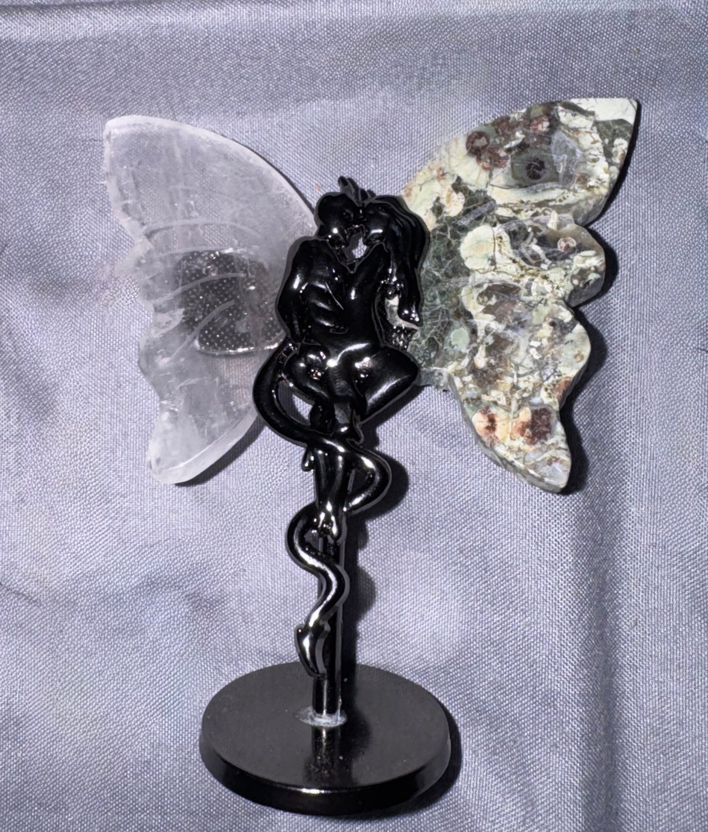 A winged  figurine with a black metallic snake-wrapped body, a clear translucent left wing, and a mottled gray-green right wing, displayed on a round black base against a gray fabric backdrop.