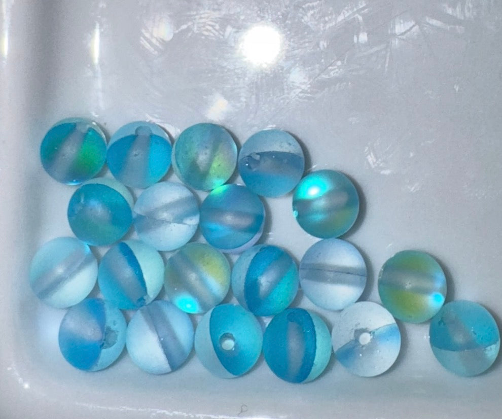 A group of round glass beads with marbled swirls of light blue, medium blue, white, and hints of green, arranged on a white surface.