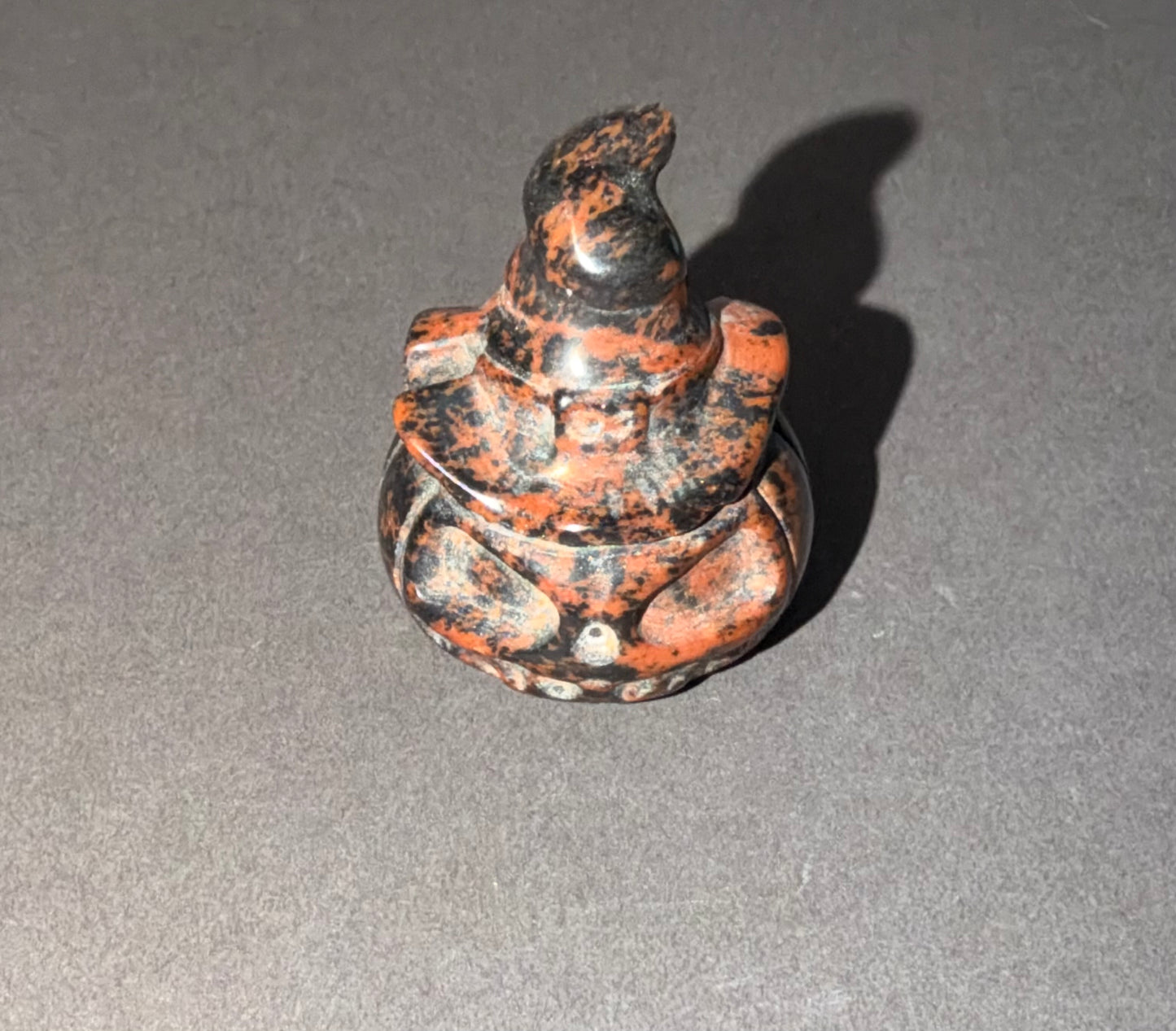 A small carved figurine of a seated figure with a pointed hat, made from a marbled stone with black and rust-orange patterns, photographed on a gray surface with a hard shadow.