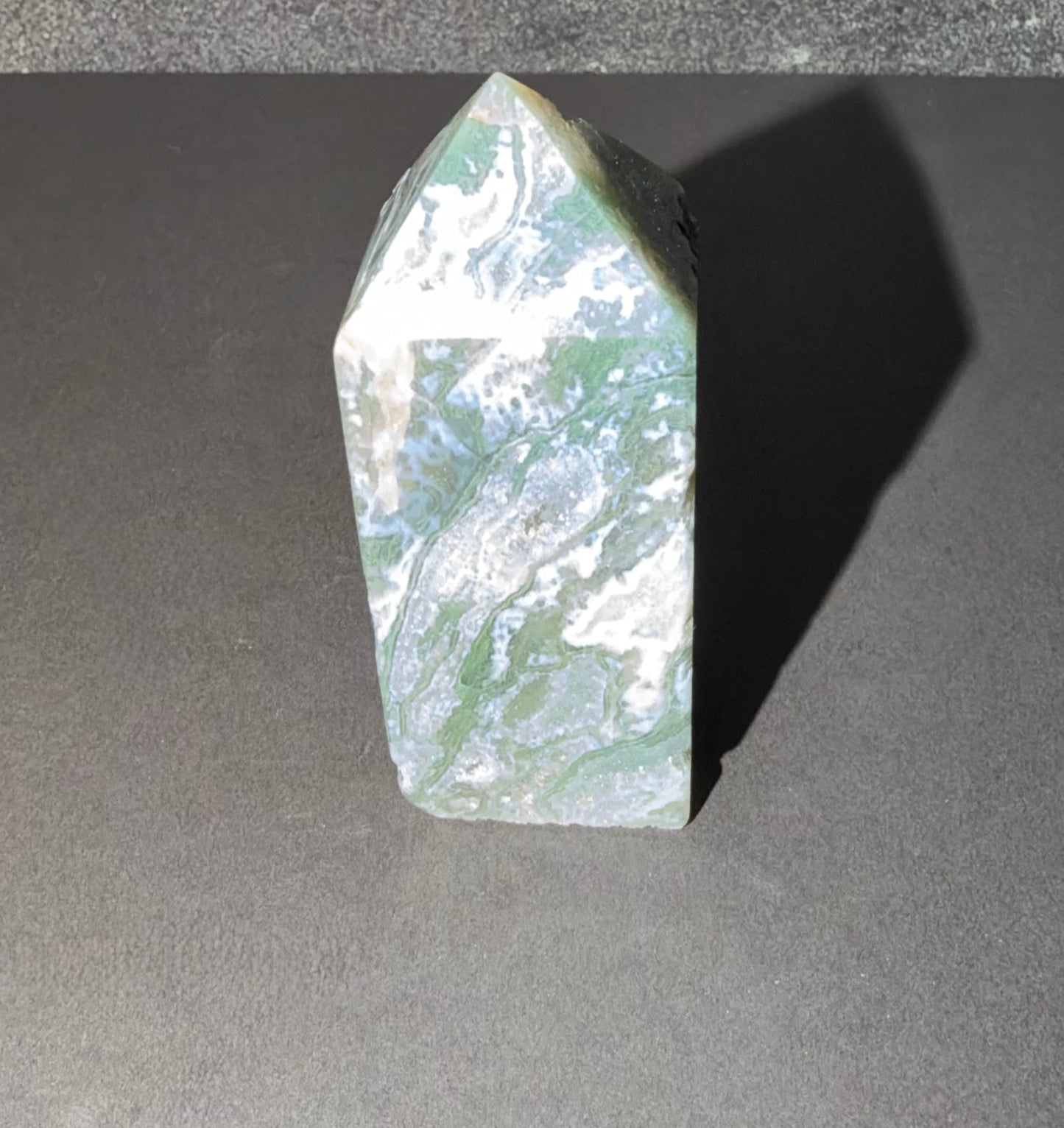 A polished natural stone tower or point with a marbled pattern of green, white, and gray, standing upright on a dark gray surface.