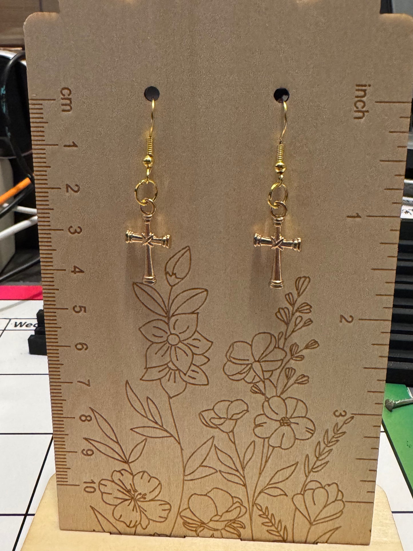 A pair of small gold cross dangle earrings displayed on a wooden measurement card with floral line art and centimeter/inch rulers.