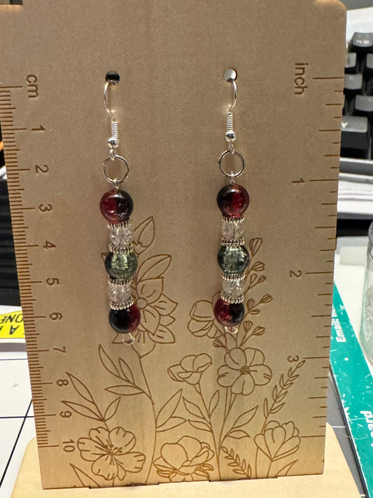 A pair of beaded dangle earrings displayed on a wooden card with engraved measurements and floral lines. Each earring has a silver fishhook, a small silver ring, a deep red bead, a green marbled bead, a clear faceted bead, and a deep red bead at the bottom.