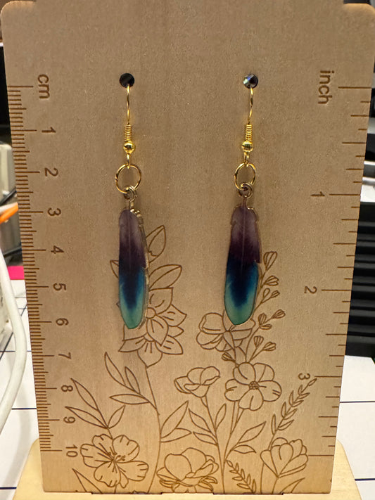 A pair of feather-shaped dangle earrings displayed on a wooden measuring board with floral line art and printed centimeter/inch rulers. The earrings have gold-tone hooks and rings with a feather design that fades from dark purple at the top to bright teal-blue at the bottom.