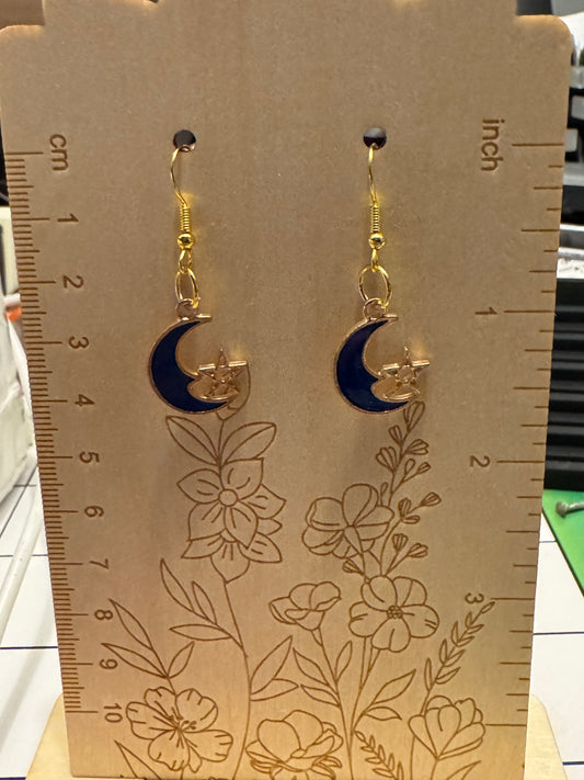 A pair of gold-toned dangle earrings with dark blue crescent moon and star charms, displayed on a wooden measuring card with centimeter and inch markings.