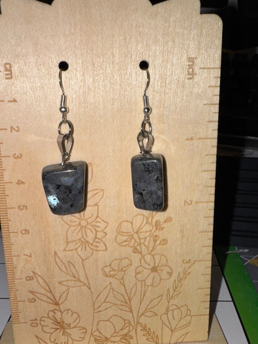 A pair of handmade dangle earrings with dark gray stone pendants and silver hooks, displayed on a wooden ruler with floral engravings.