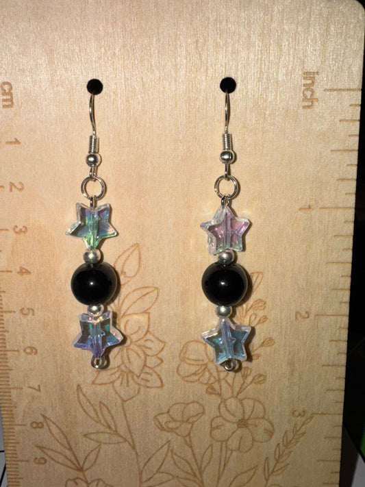 A pair of handmade dangle earrings displayed on a wooden ruler. The earrings feature silver fishhook wires, a small silver bead, a round black bead, and two iridescent star-shaped beads at the bottom.