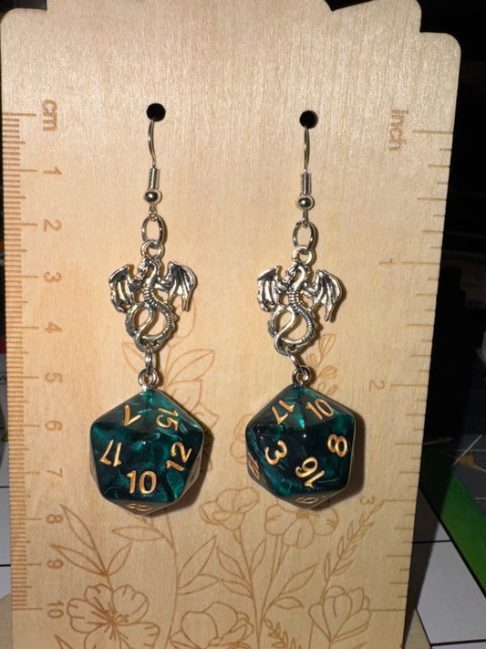 A pair of fantasy-themed d20 dice earrings hanging on a wooden display stand. Each earring features a silver dragon charm and a dangling green marbled polyhedral die with gold numbers.