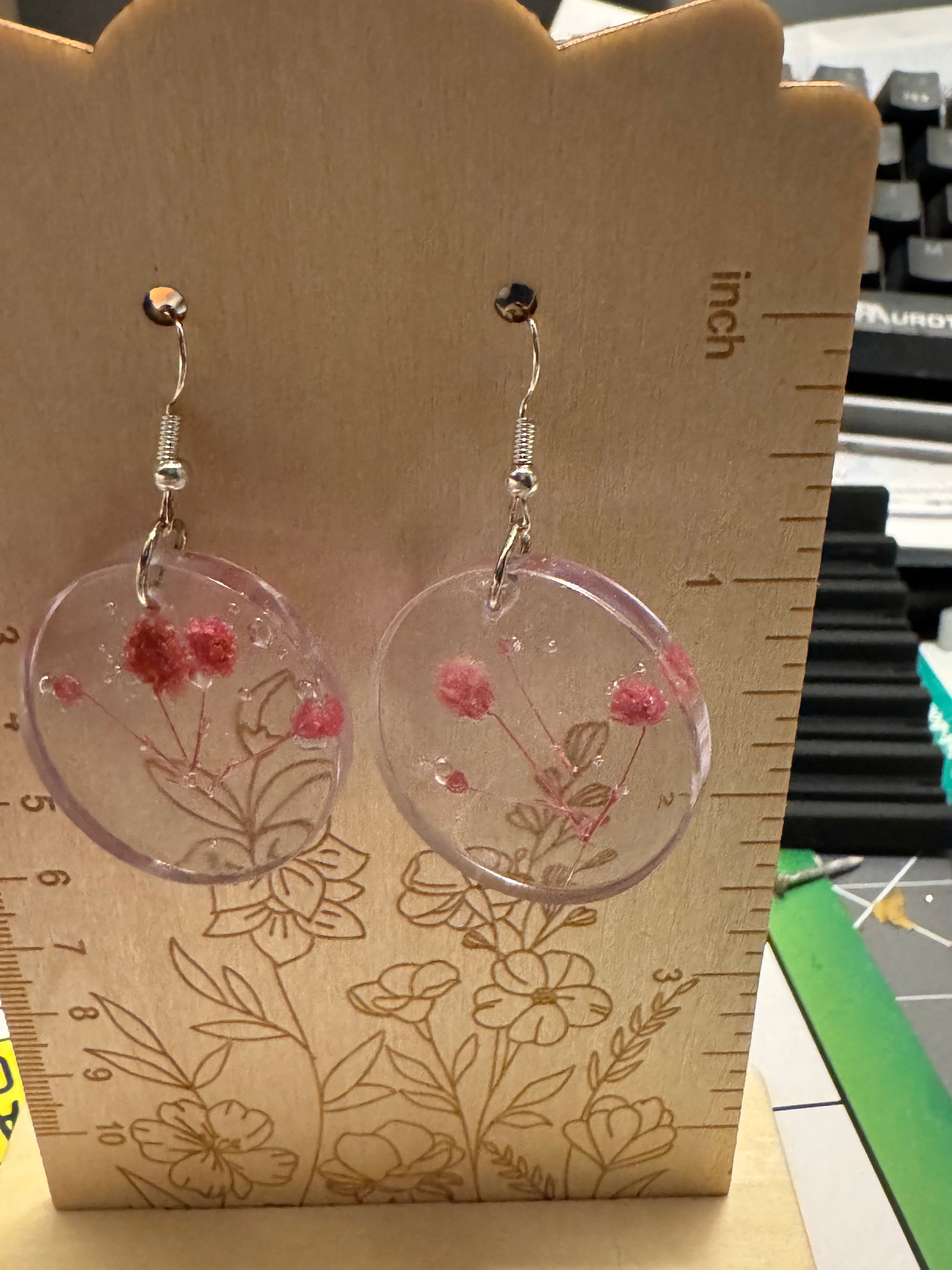 A pair of round clear resin drop earrings with embedded pink dried flowers, hanging on silver hooks from a wooden display block with inch and floral measurements.