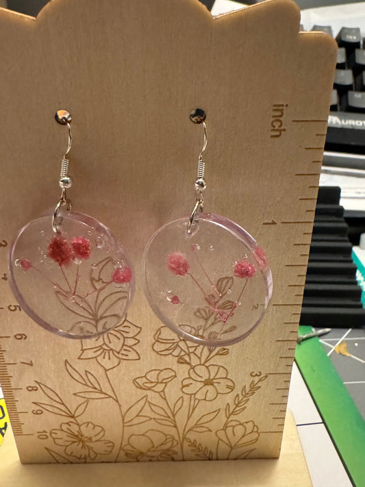 A pair of round clear resin drop earrings with embedded pink dried flowers, hanging on silver hooks from a wooden display block with inch and floral measurements.