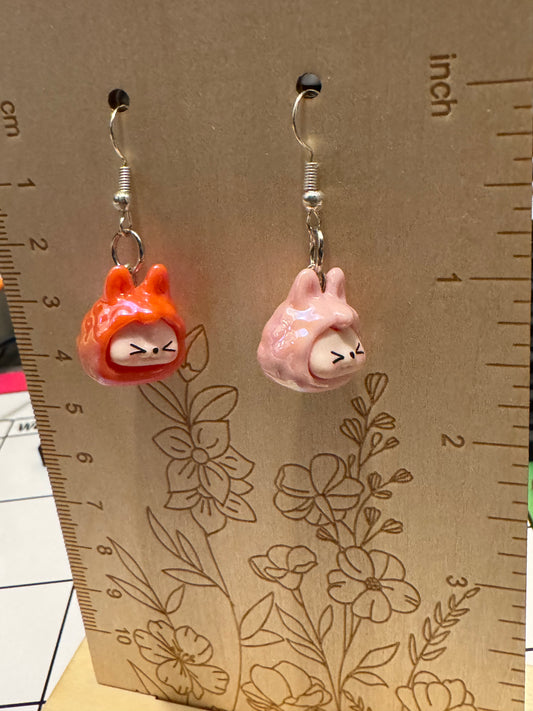 Two small dangling earrings displayed on a wooden measuring card. One earring has an orange-red hooded figure with a white face, and the other has a pink rabbit-like figure with a white face.