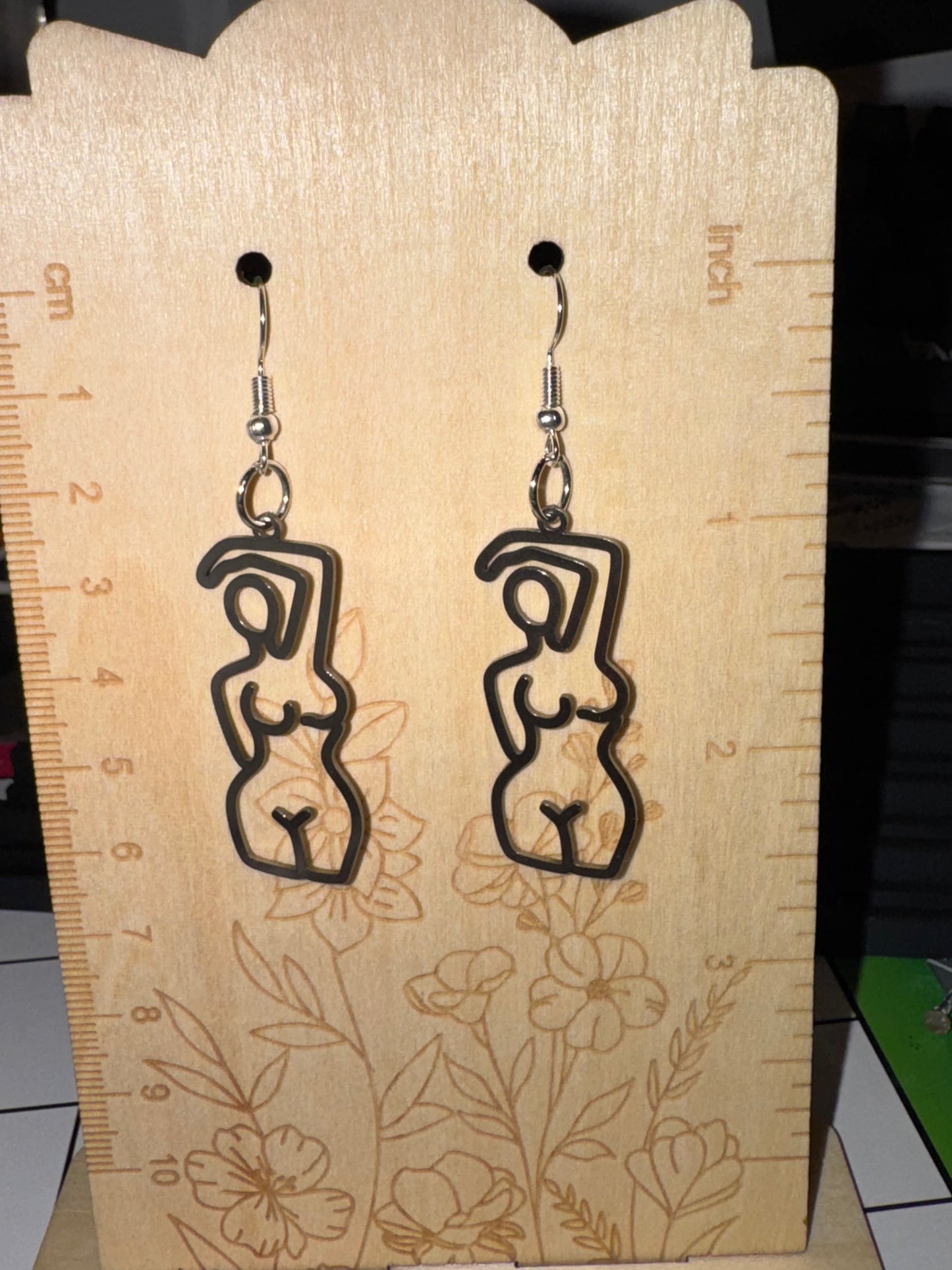 A pair of black laser-cut dangle earrings shaped like abstract female figures, displayed on a wooden ruler stand.