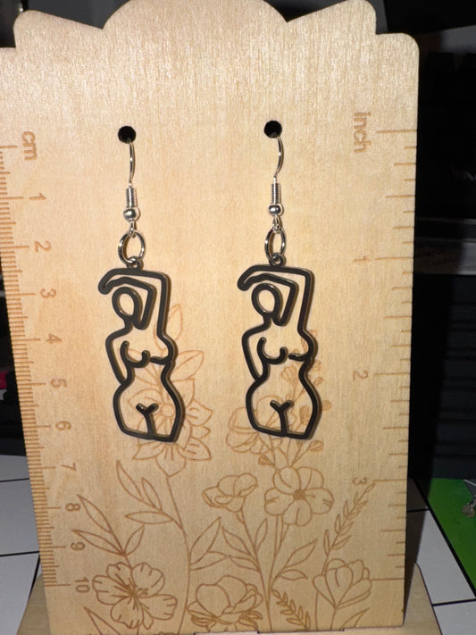 A pair of black laser-cut dangle earrings shaped like abstract female figures, displayed on a wooden ruler stand.