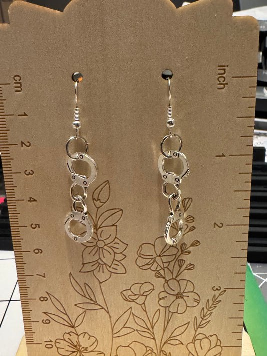 A pair of silver-toned dangle earrings displayed on a brown kraft card with engraved floral lines and printed inch/centimeter rulers along the left and right edges. The earrings feature stacked circular metal rings and small clear rhinestone accents.