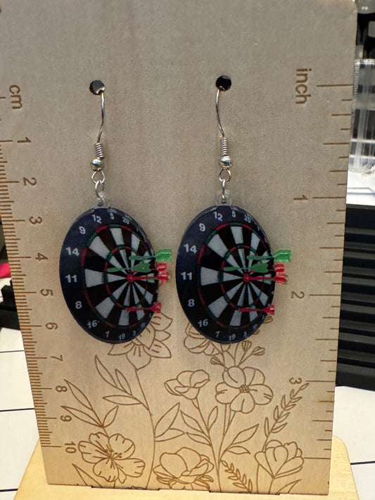 A pair of dangling earrings shaped like dartboards, hanging on a tan display card with printed measurements and floral line art. The earrings have black, white, gray, red, green, and yellow dartboard details with silver hooks.