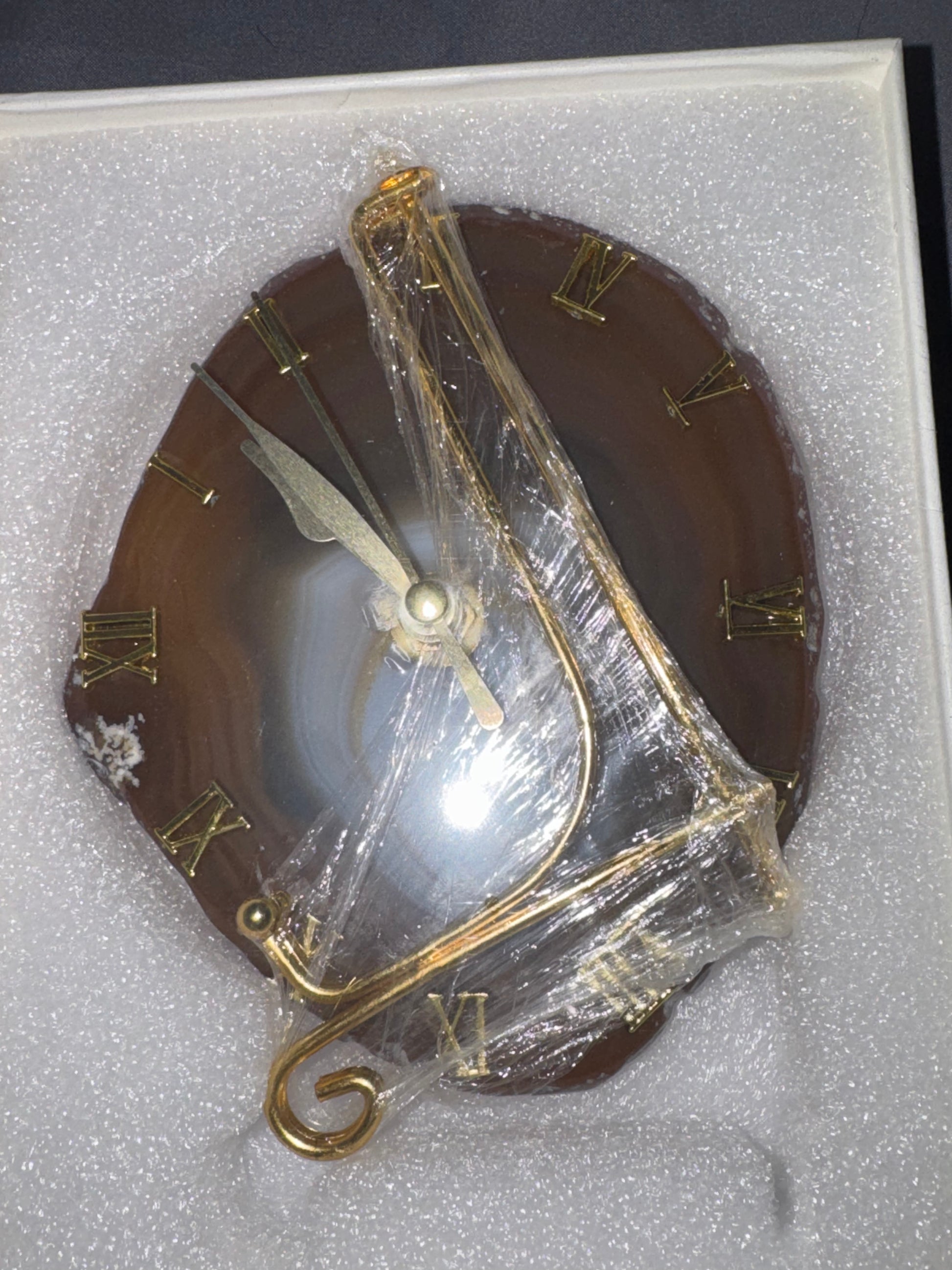 A round brown agate clock face with gold Roman numerals and gold-toned hands, wrapped in clear protective plastic inside a white foam display box.