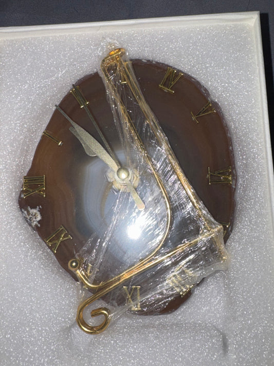 A round brown agate clock face with gold Roman numerals and gold-toned hands, wrapped in clear protective plastic inside a white foam display box.
