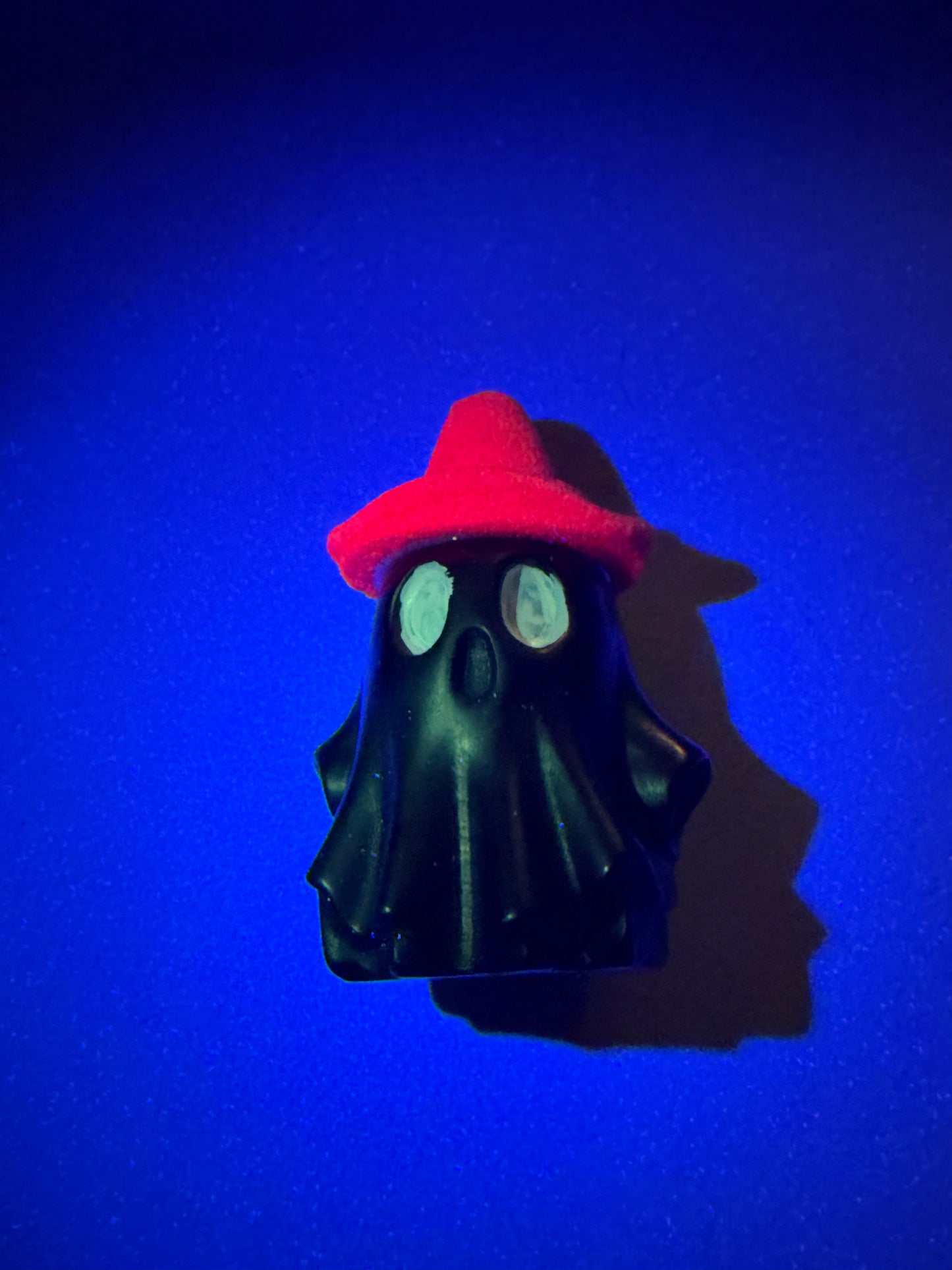 A small black ghost-shaped accessory with glowing white eyes and a bright red hat, photographed against a vivid blue background with a strong shadow.