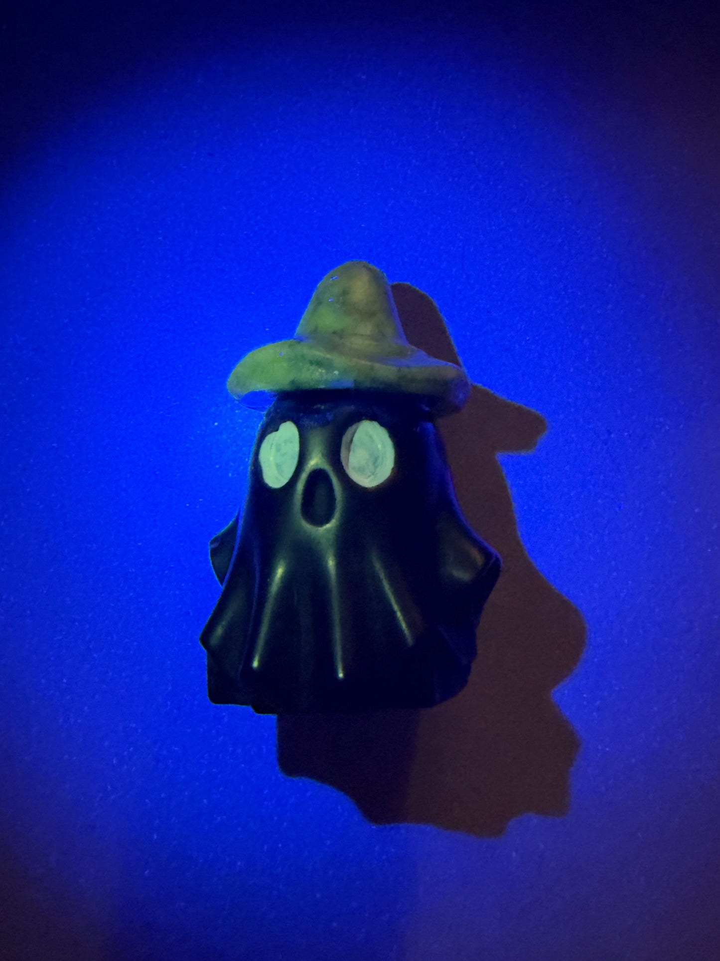 A small black ghost-shaped figurine wearing a green hat, photographed against a dark blue background with a strong shadow cast to the right.