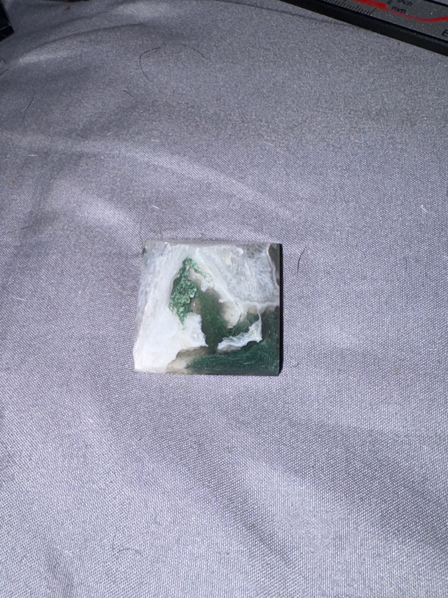 A small square stone or tile with a marbled green and white pattern, placed on a gray textured fabric surface.