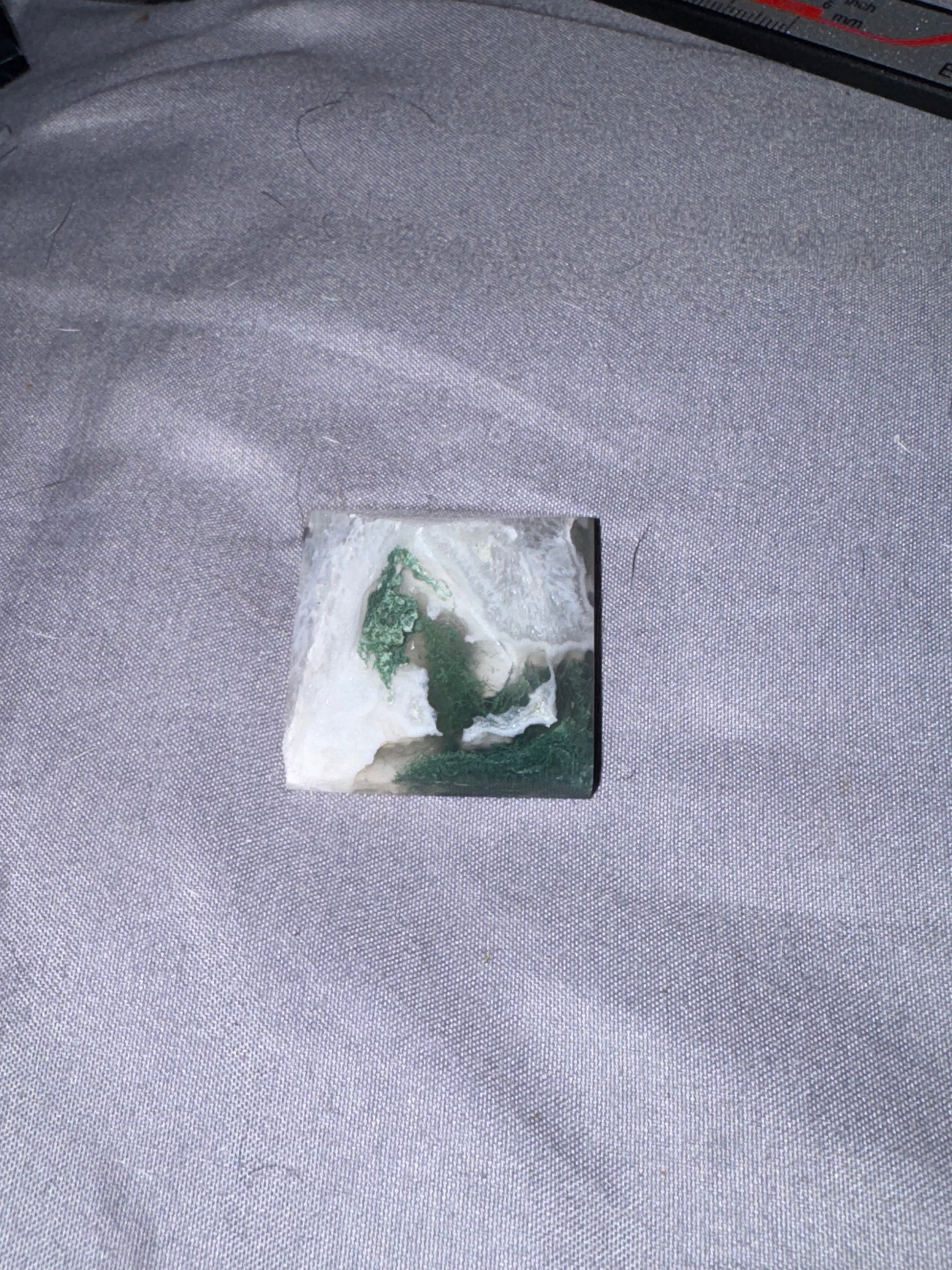 A small square stone or tile with a marbled green and white pattern, placed on a gray textured fabric surface.