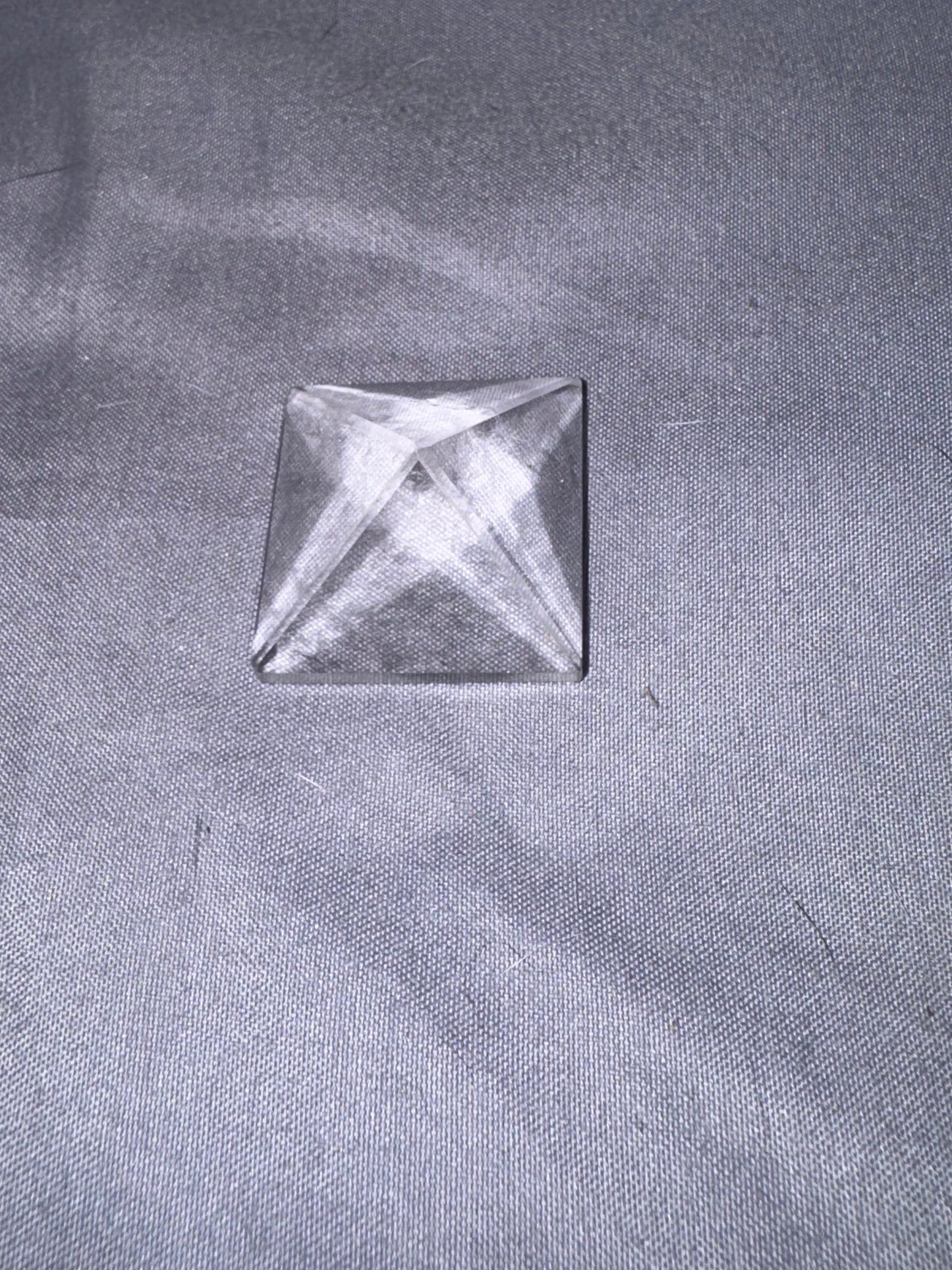 A clear glass pyramid cabochon with a square base, resting on a textured gray fabric background.
