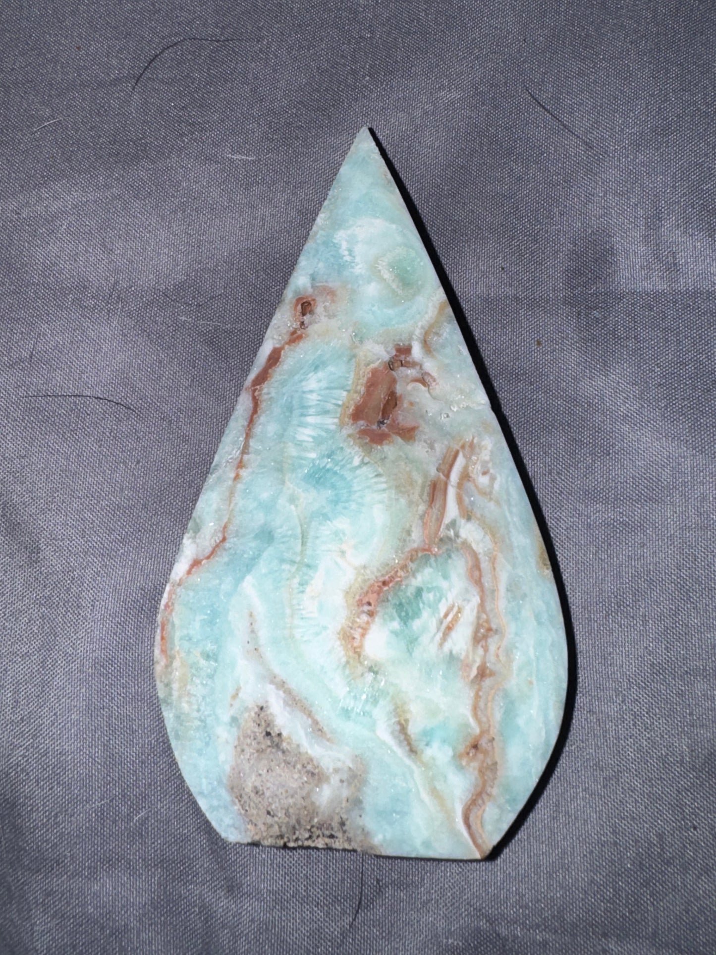 A polished teardrop-shaped stone slab with natural marbling in light blue, white, and brown, displayed on a textured dark gray fabric background.